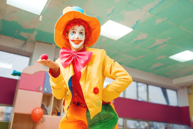 Payaso divertido posa con nariz roja en la mano. Fiesta de cumpleaños celebrando en la sala de juegos, vacaciones para bebés en el patio de recreo