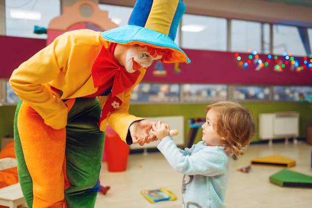 Payaso divertido reparte piruletas a la niña feliz, amistad para siempre. Fiesta de cumpleaños celebrando en la sala de juegos, vacaciones para bebés en el patio de recreo.