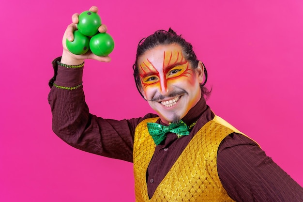 Foto payaso con maquillaje facial blanco sobre un fondo rosa haciendo malabares con bolas verdes