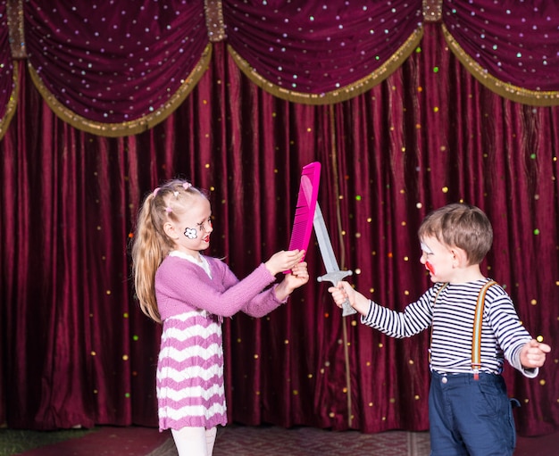 Payasos jóvenes con pelea de espadas en el escenario con accesorios, niña sosteniendo un gran peine rosa mientras que el niño sostiene la espada de juguete