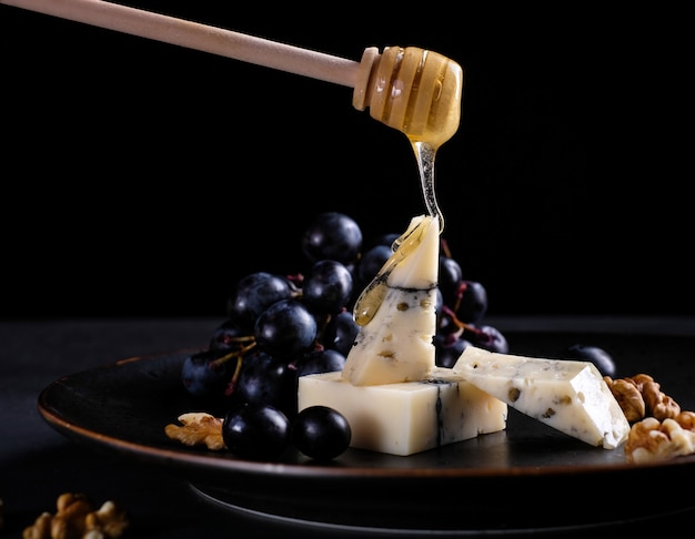 Paz de queso hecho a mano en un plato con uvas, nueces y miel en mesa negro. De cerca.