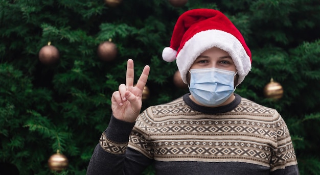 Peça de Natal. Feche o retrato de um homem com um chapéu de Papai Noel, uma camisola de Natal e uma máscara médica com emoção. No contexto de uma árvore de Natal. Pandemia do coronavírus