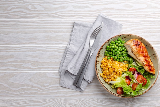 Pechuga de pollo al horno en un plato con ensalada fresca, guisantes y maíz, fondo de madera blanca, vista superior. Comida fitness saludable con filete de pollo, equilibrada en proteínas y carbohidratos. Arriba, copie el espacio