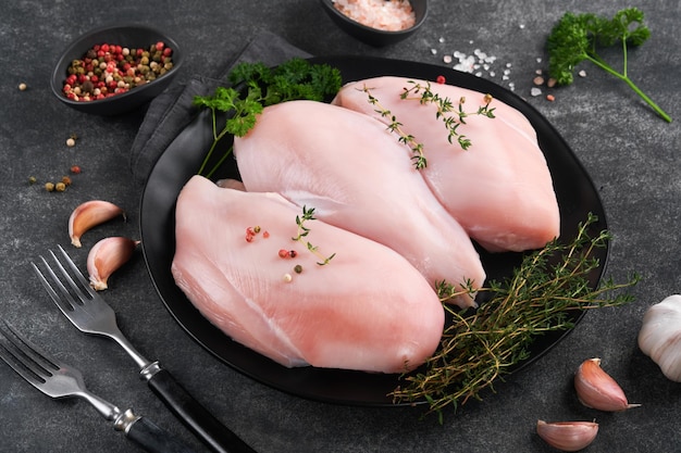 Pechuga de pollo Filetes de pechuga de pollo cruda en placa de cerámica negra sobre tabla de cortar de madera con hierbas y especias sobre fondo negro Vista superior con espacio de copia