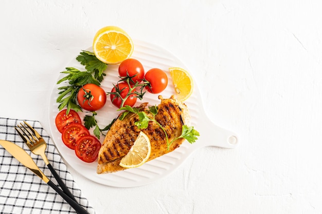 Pechuga de pollo a la parrilla en diferentes variaciones con tomates cherry, hierbas verdes, limón cortado en una bandeja de cerámica blanca Vista superior