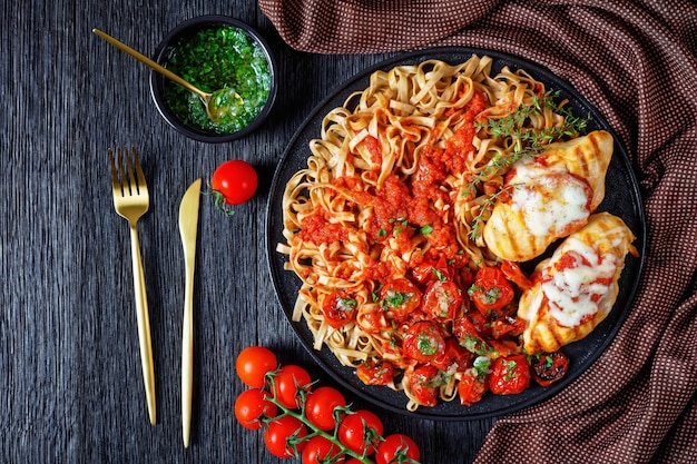 Pechuga de pollo a la parrilla con linguini de champiñones con salsa de tomate, tomates cherry al horno cubiertos con salsa de ajo, servido con tomillo en un plato negro sobre una mesa de madera, vista superior, endecha plana