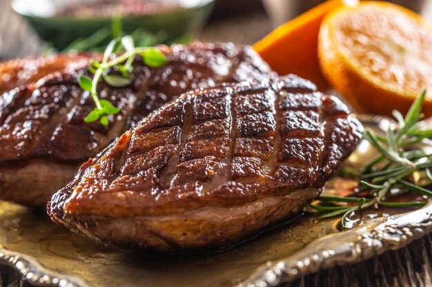 Pechugas de pato recién asadas descansando sobre un plato con hierbas y naranjas.