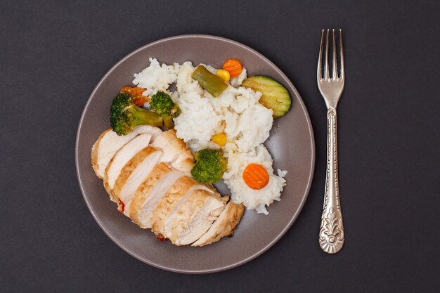 Pechugas de pollo al horno o filete con arroz, verduras y verduras en un plato con un tenedor. Fondo negro. Vista superior.