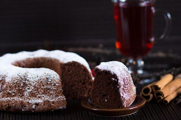 Pedaço de bolo de chocolate de natal com açúcar em pó na mesa de madeira com copo de chá vermelho e paus de canela