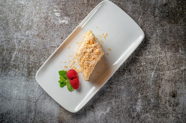 Pedaço de bolo de creme em camadas com framboesas e hortelã em um prato branco contra uma mesa de pedra cinza em um restaurante, um delicioso café da manhã