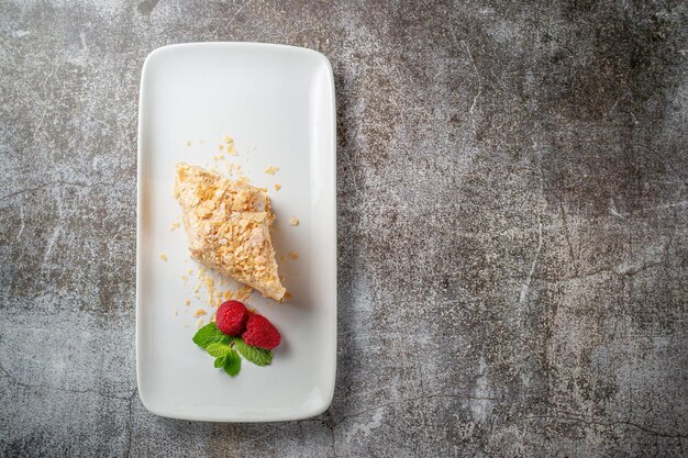 Pedaço de bolo de creme em camadas com framboesas e hortelã em um prato branco contra uma mesa de pedra cinza em um restaurante, um delicioso café da manhã