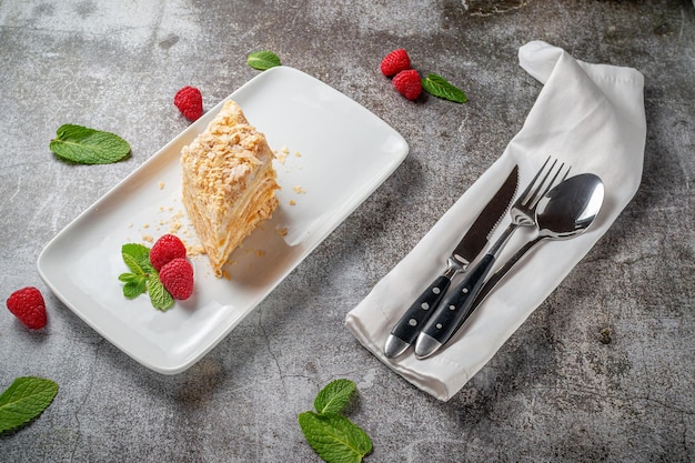 Pedaço de bolo de creme em camadas com framboesas e hortelã em um prato branco contra uma mesa de pedra cinza em um restaurante, um delicioso café da manhã