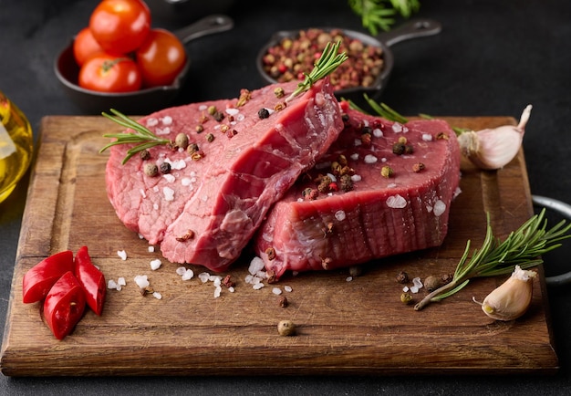 Pedaço de carne crua com especiarias pimenta raminho de alecrim sal e azeite em uma placa de madeira fundo preto