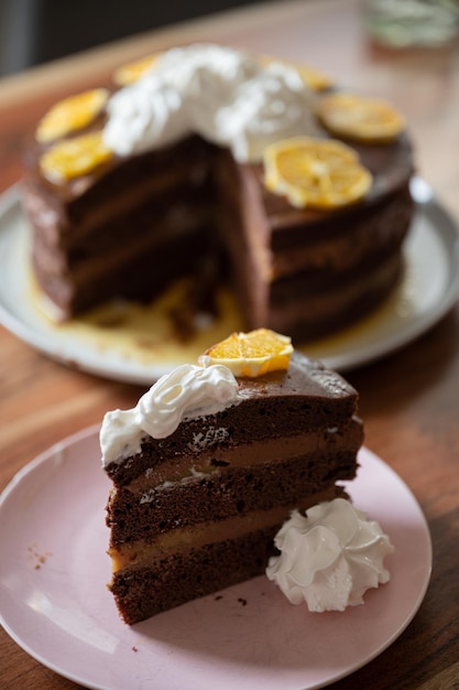 Pedaço de delicioso bolo de chocolate laranja vegano servido com chantilly em um prato com o bolo inteiro ao fundo