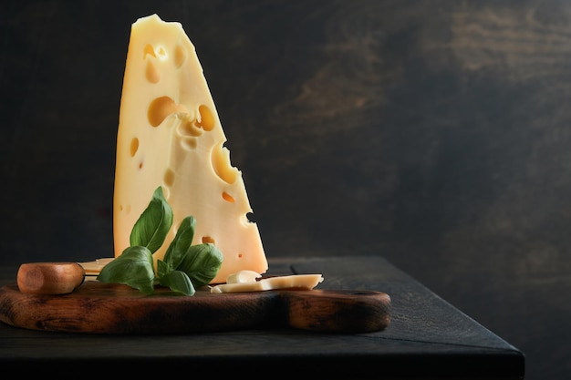 Pedaço de queijo de triângulo de queijo Queijo suíço com manjericão e uma faca em uma tábua de servir no fundo da mesa de madeira escura Fundo de queijo