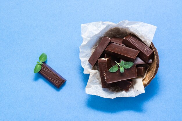 Pedaços de chocolate e folhas de hortelã em uma tigela e sobre fundo de papel azul. O conceito de excesso de peso de doces. Vista do topo