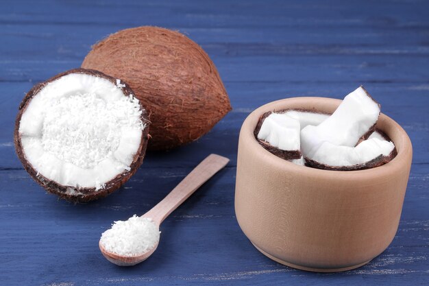 Pedaços de coco em uma tigela de madeira com uma colher com um grande coco inteiro sobre um fundo azul de madeira.