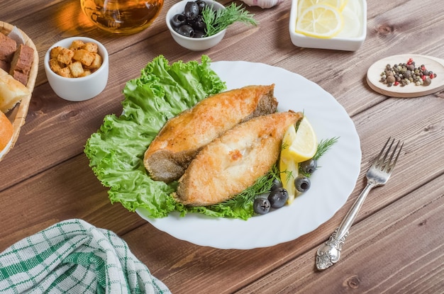 Pedaços de filé de peixe frito com limão e ervas em um prato branco sobre um fundo escuro de madeira.