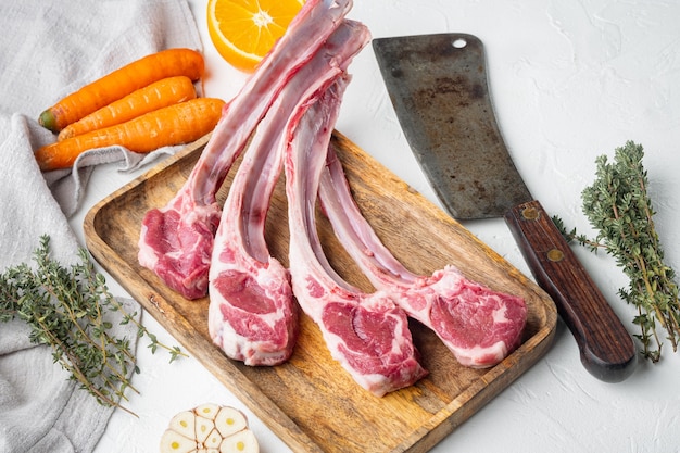 Pedaços orgânicos de carne de carneiro, Costeleta de cordeiro, crua com osso definido, com ingredientes laranja cenoura, ervas e faca de cutelo de açougueiro velha, em mesa de pedra branca