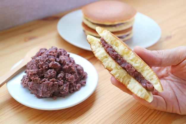 Pedazo de Half Dorayaki en la mano con un plato de pasta de frijol rojo sobre mesa de madera