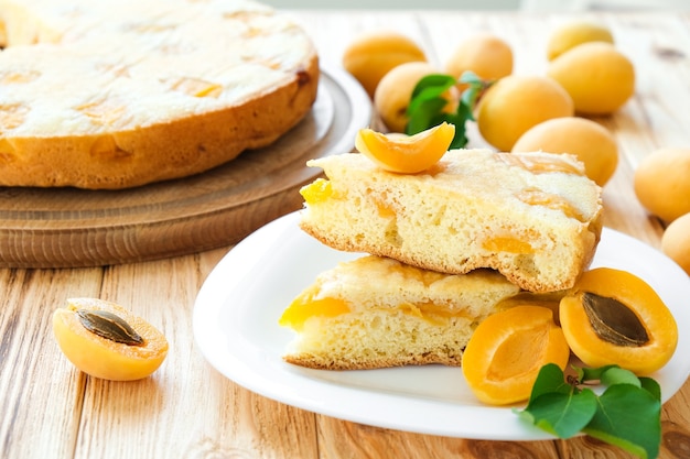 Pedazo de pastel de albaricoque en un plato con pastel completo y frutas de albaricoque en el fondo de la tabla de madera. Receta fácil de merienda navideña. Panadería casera por receta clásica. Postre vegano, sin gluten, sin azúcar.