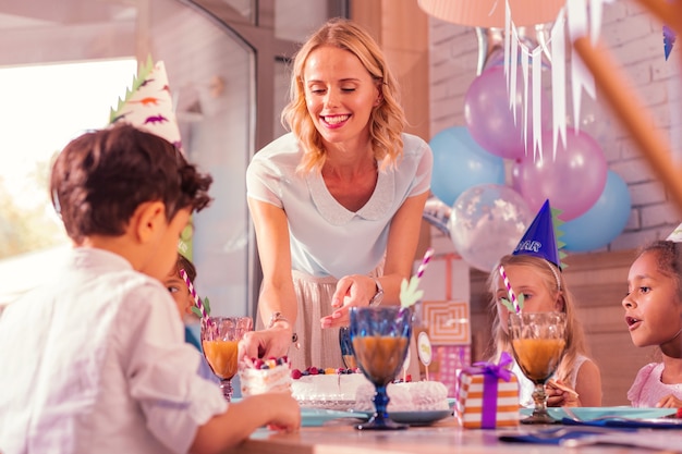 Pedazo de pastel. Mujer joven alegre sonriendo y dando un pedazo de pastel al niño en la fiesta de cumpleaños