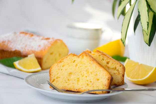 Pedazo de tarta de limón en un plato sobre una mesa de mármol y una planta verde en una maceta con tarta completa y limones en el fondo. Panadería casera por receta clásica. Delicioso postre para la hora del té del desayuno.