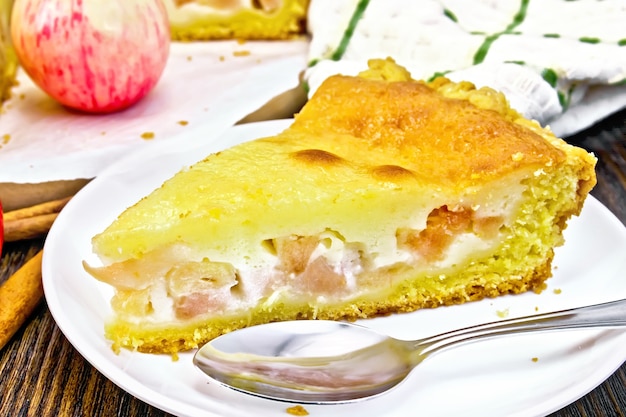 Pedazo de tarta de manzana con salsa de crema agria en un plato blanco, canela, fruta y servilleta sobre un fondo de una tabla de madera oscura.