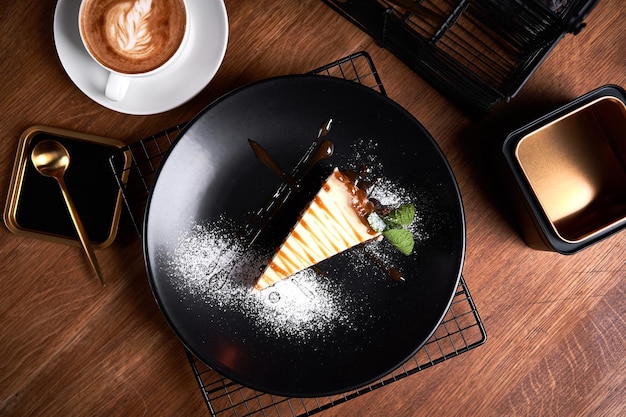 Pedazo de tarta de queso en un plato negro sobre una mesa con menta y una taza de café Vista lateral