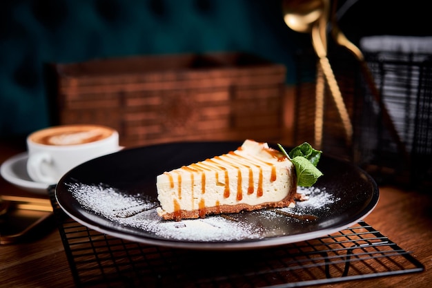 Foto pedazo de tarta de queso en un plato negro sobre una mesa con menta y una taza de café vista lateral