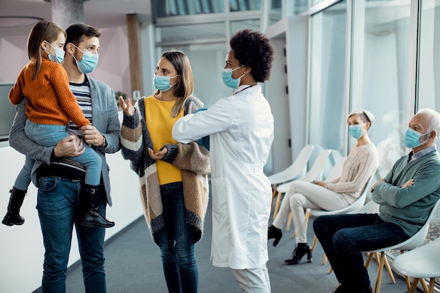 Pediatra afro-americano conversando com uma jovem família em um corredor na clínica médica durante a pandemia de coronavírus