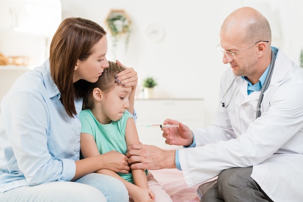 Pediatra contemporâneo injetando em uma menina sentada ao lado da mãe no consultório médico