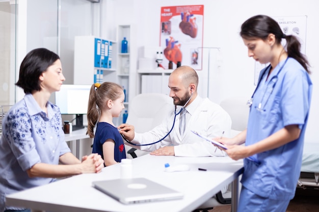 Pediatra escuchando corazón de niño enfermo con estetoscopio en la oficina del hospital y enfermera tomando notas. Médico especialista en medicina que brinda servicios de atención médica. Examen de tratamiento.