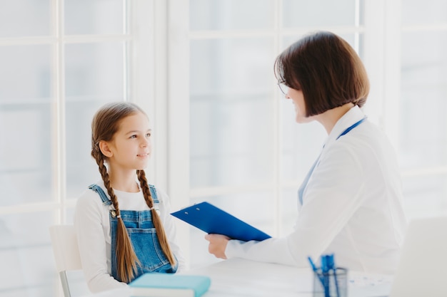 Pediatra examina criança pequena, ouve atentamente o paciente criança pequena, escreve notas na área de transferência