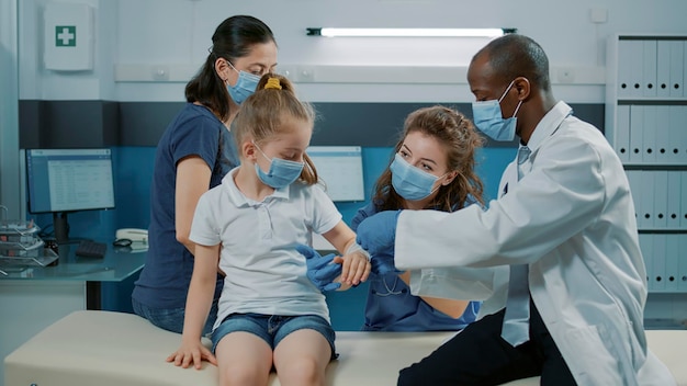 Pediatra poniendo tirita en el brazo lesionado del niño en consulta en el gabinete. Médico masculino que examina la fractura para ayudar a la niña con la recuperación en la cita médica durante la pandemia.