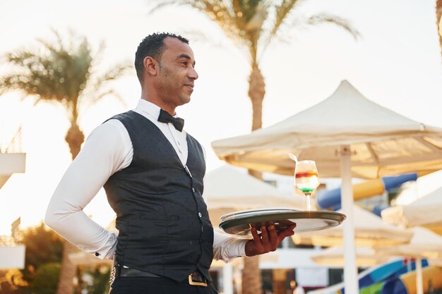 Con el pedido en la mano, el camarero negro con ropa formal está en su trabajo al aire libre durante el día soleado