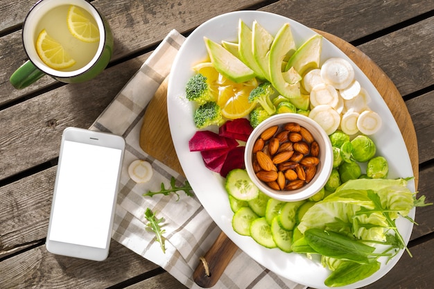 Pedidos de comida en línea. Plato saludable comida vegana con smartphone con pantalla en blanco en la vista superior de la mesa de madera