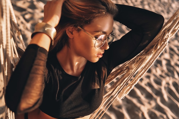 Pegando o sol. Vista superior de uma jovem atraente de óculos, mantendo as mãos no cabelo enquanto está sentado em uma rede na praia
