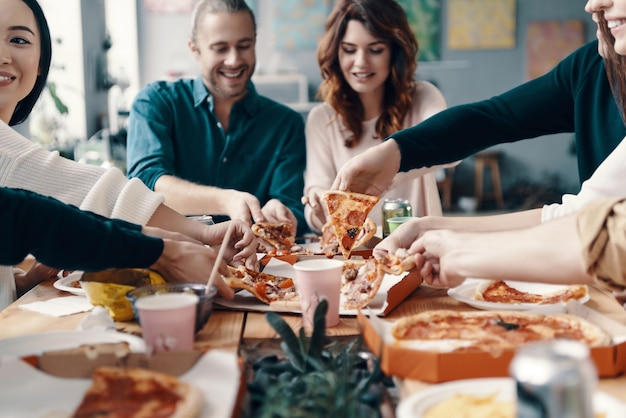 Pegando outra fatia. grupo de jovens em trajes casuais pegando pizza e sorrindo durante um jantar dentro de casa