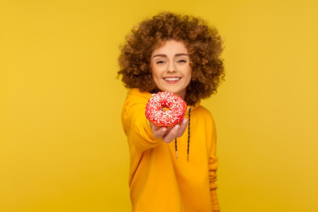 Pegue a sobremesa doce. Retrato de uma jovem de cabelos cacheados feliz com capuz estilo urbano dando delicioso donut açucarado e sorrindo genuinamente para a câmera. tiro de estúdio interno isolado em fundo amarelo