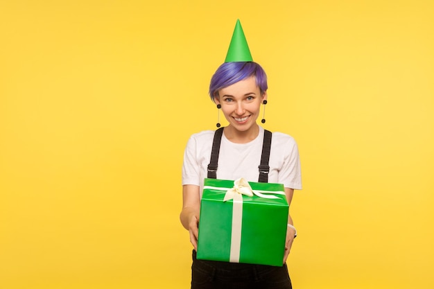 Pegue o presente de aniversário retrato de uma generosa garota hipster fofa com cabelo curto violeta usando cone de festa engraçado e macacão dando caixa de presente parabenizando isolado em fundo amarelo studio shot