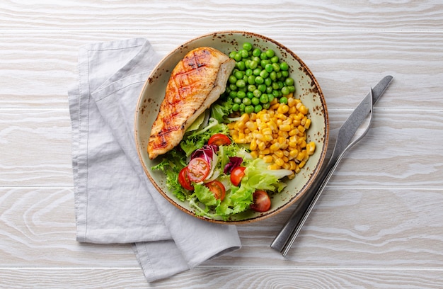 Foto peito de frango assado no prato com salada fresca, ervilhas e milho, fundo branco de madeira, vista superior. refeição saudável de fitness com filé de frango, balanceada em proteínas e carboidratos. dieta e nutrição