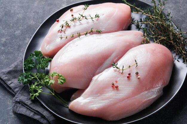 Peito de frango Filetes de peito de frango cru na placa de cerâmica preta na tábua de madeira com ervas e especiarias em fundo preto Vista superior com espaço de cópia