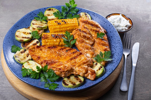 Peito de frango grelhado com milho e abobrinha em um prato azul close-up. Prato de verão com frango grelhado e legumes.