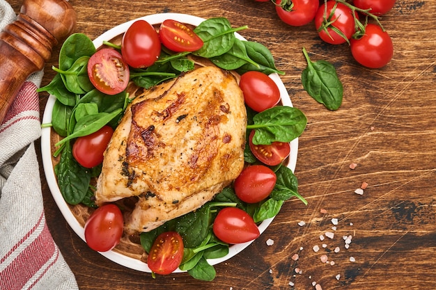 Peito de frango grelhado com salada verde de espinafre, pimenta e tomate cereja em uma placa de cerâmica em um fundo de mesa de madeira velha. Vista do topo.