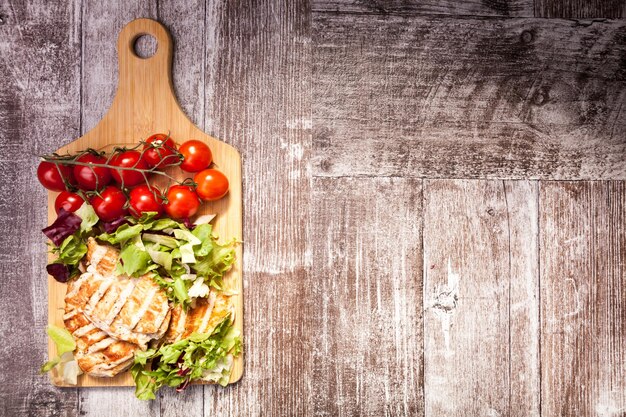 Peito de frango grelhado em salada recém-cortada ao lado de um cacho de tomates em uma placa de madeira