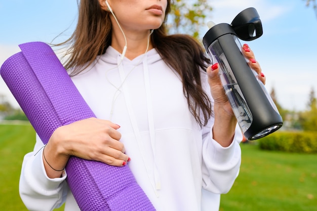 Peito de menina esportiva com capuz branco segura tapete roxo, garrafa de água, usando fones de ouvido após o treino, exercício físico ao ar livre