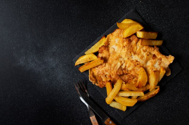 Foto peixe britânico tradicional com batatas fritas. em uma placa preta e um fundo preto. espaço para texto. vista superior, plana leigos.