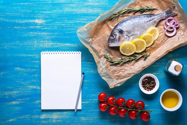 Foto peixe dourado em papel de cozinha com especiarias e um caderno de receita ou menu. vista superior sobre um fundo azul.