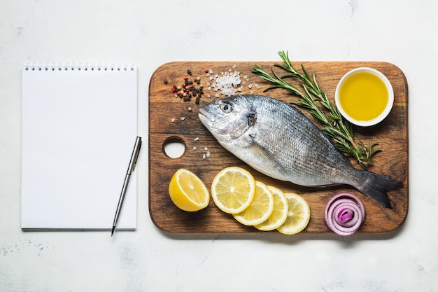 Peixe dourado em uma tábua de madeira rústica com especiarias e um caderno de receita ou menu. Vista superior em um fundo branco.